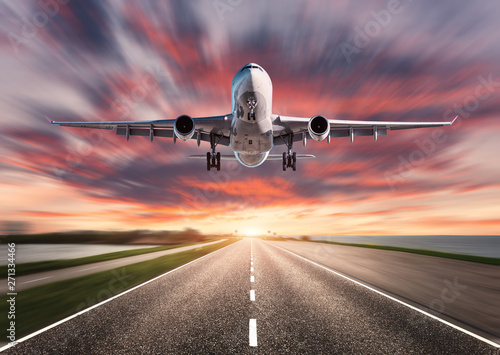 Airplane and road with motion blur effect at sunset. Landscape with passenger airplane is flying over asphalt road and colorful sky. Commercial plane is landing. Aircraft with blurred background
