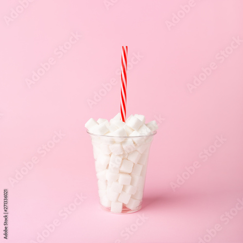 Plastic cup with straw full of sugar cubes on pastel pink background. Unhealthy food concept. Minimal, vertical, side view.