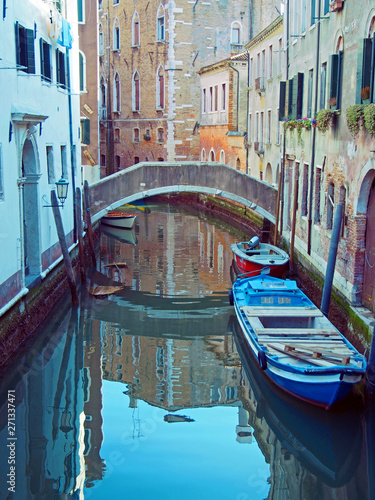 a quiet narrow canal crossed by a bridge with moored boats and old buildings reflected in the water in Venice