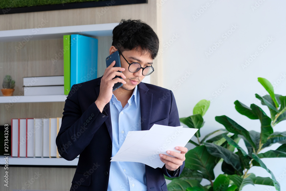 Young asian businessman reading papers talking phone at office, business communication and technology concept Young asian businessman reading papers talking phone at office, business communication and technology concept