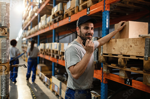 Happy warehouse worker talking on walkie-talkie in a storage room. © Drazen