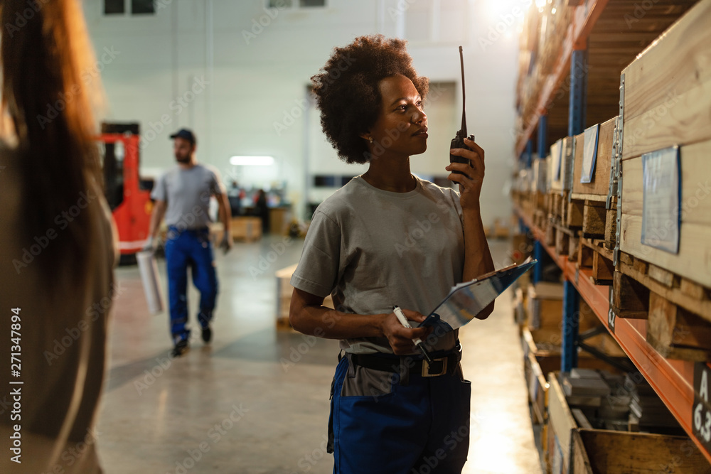 African American female dispatcher communicating on walkie-talkie in distribution warehouse.
