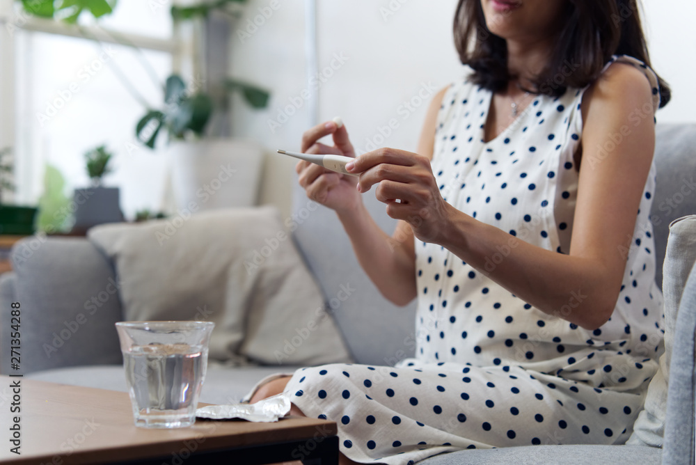 Sickness or illness of pregnancy woman concept. Asian pregnancy girl has a fever, she reading temperature on thermometer and taking a pill of medicine sitting on sofa at home. Sickness or illness of pregnancy woman concept. Asian pregnancy girl has a fever, she reading temperature on thermometer and taking a pill of medicine sitting on sofa at home.