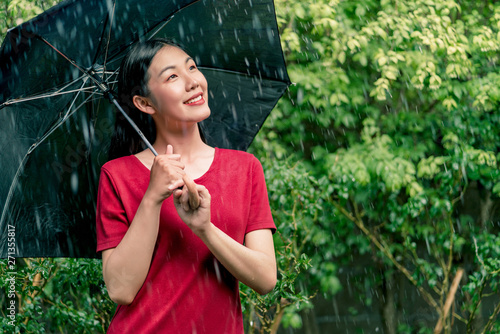 Young asian woman wearing red T-shirt smiling under black umbrella in the rain, Green nature background, Rainy season.