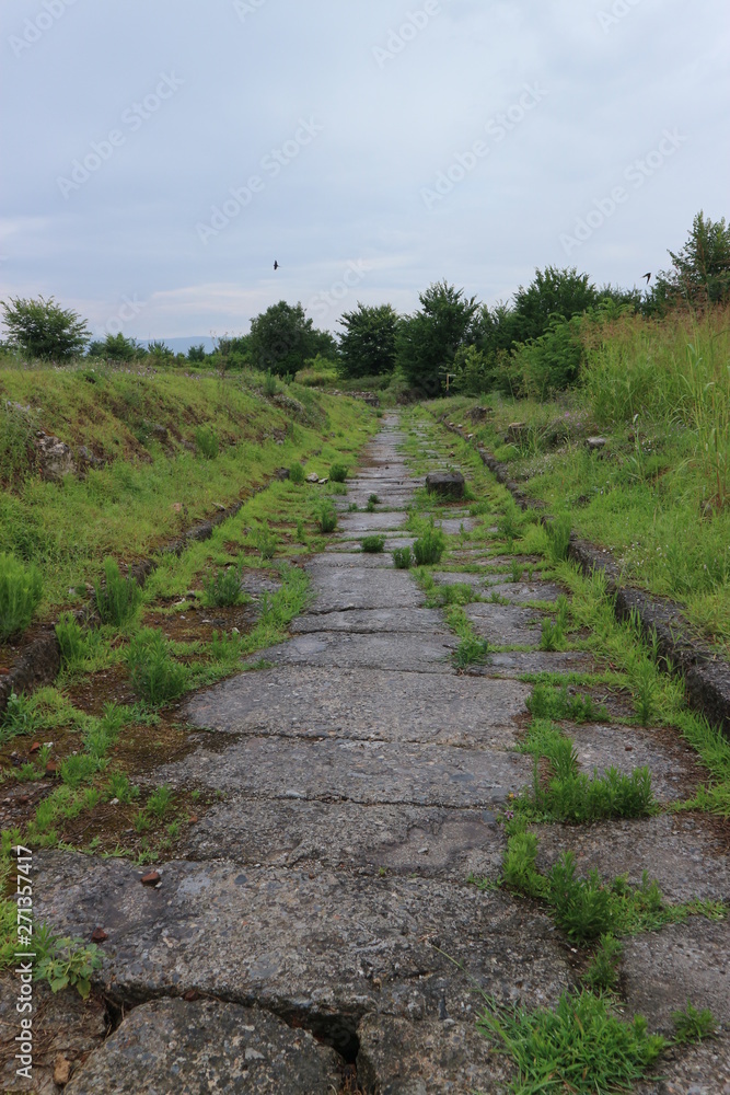 Obraz premium Abandoned way - ancient greek stone road surrounded by green hills, archaeological site Dion, Greece