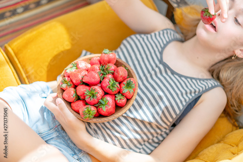 Attractive young woman laying on sofa in a home family room living room, eating fresh strawberry. Girl eating healthy food at home, interior. Well being lifestyle