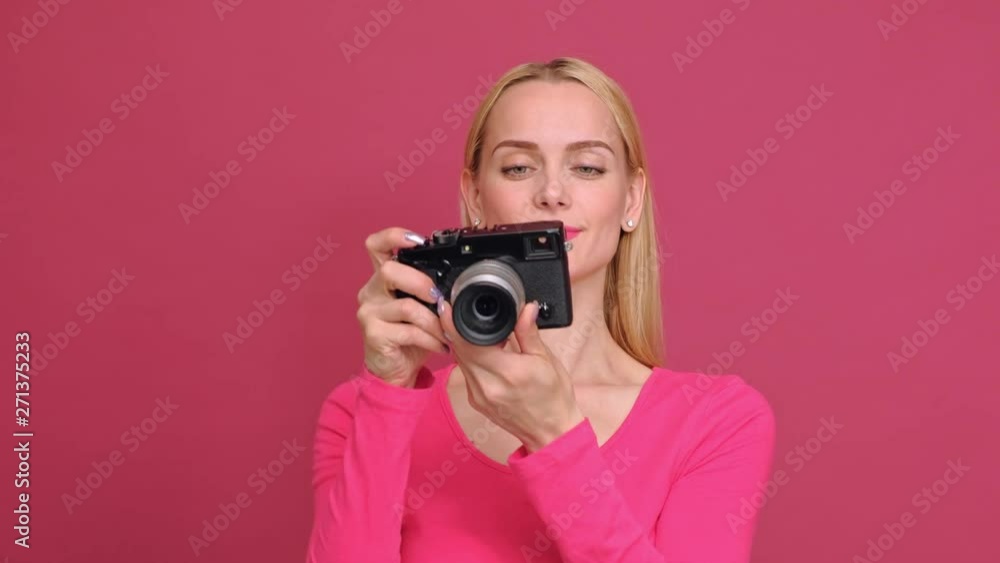 Attractive young woman blonde in a red T-shirt and jeans posing on a pink background. Photographs on a mirrorless camera in retro style and smiling. The concept of the photographer. Attractive young woman blonde in a red T-shirt and jeans posing on a pink background. Photographs on a mirrorless camera in retro style and smiling. The concept of the photographer.