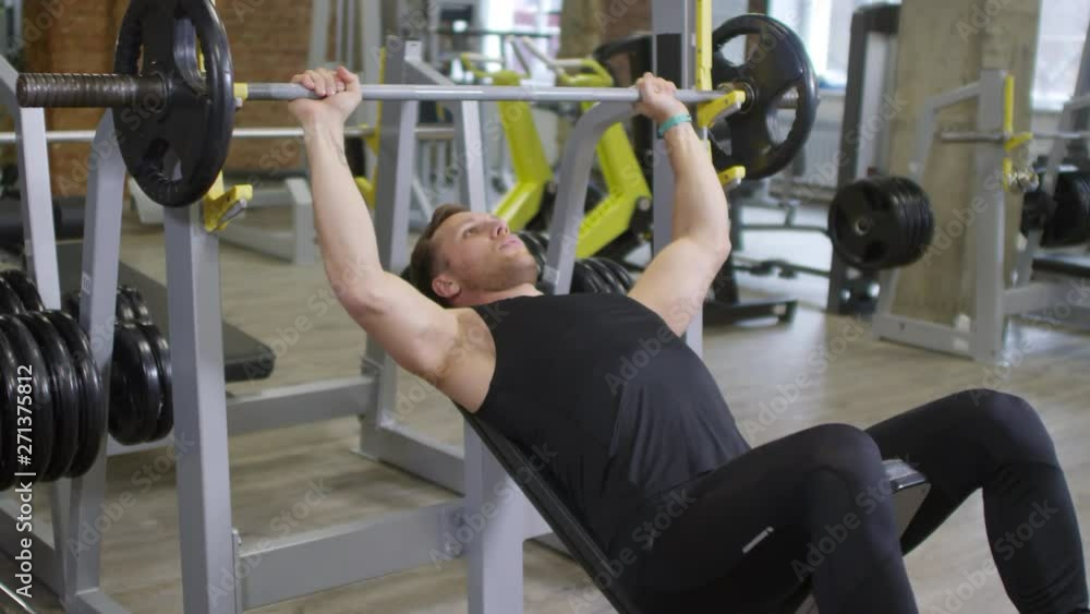 Professional male coach in sportswear speaking on camera and demonstrating barbell bench press on camera while recording online workout in gym Professional male coach in sportswear speaking on camera and demonstrating barbell bench press on camera while recording online workout in gym