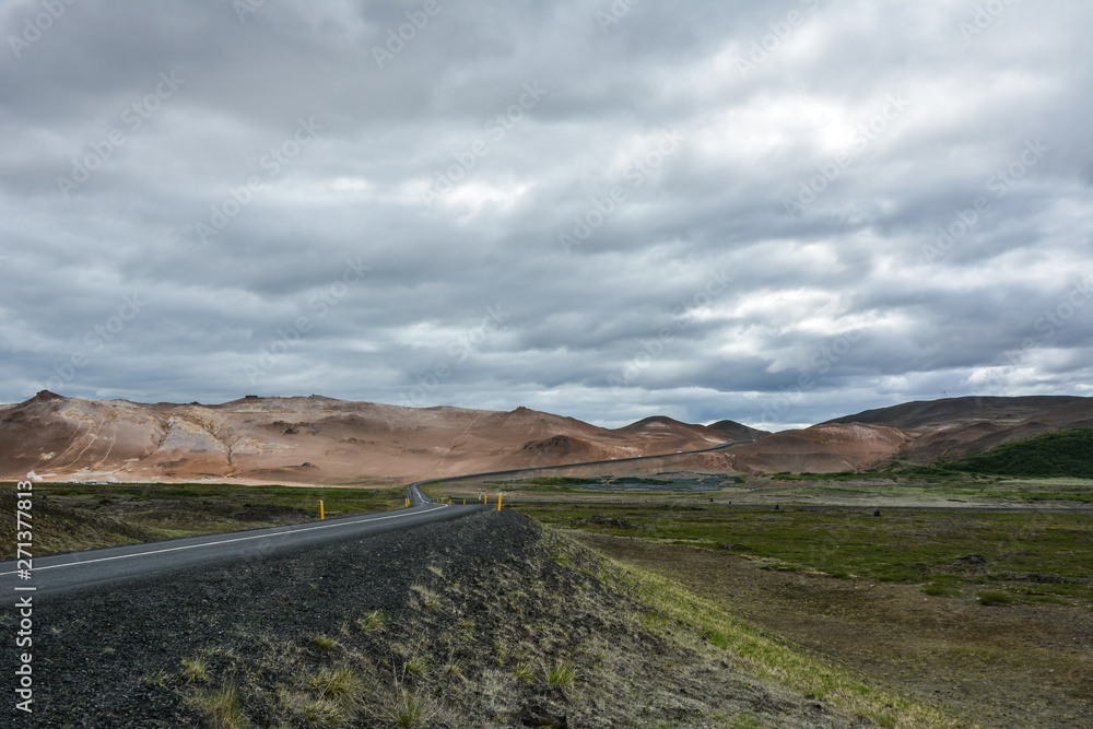 Curvy road, empty meadow and red mountains in the background in Myvatn region, overcast day in summer