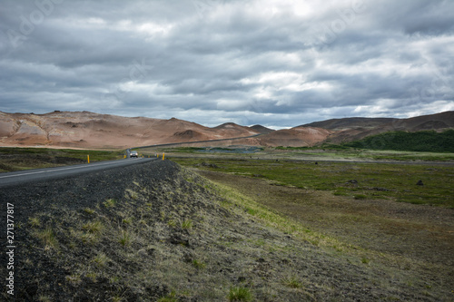 Road, empty meadow and red mountains in the background with a car in Myvatn region, overcast day in summer