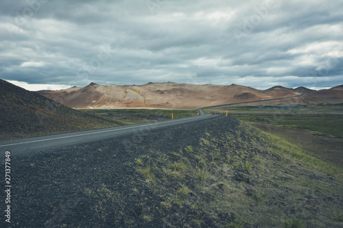 Curvy road, empty meadow and red mountains in the background in Myvatn region, overcast day in summer, vintage effect with grain