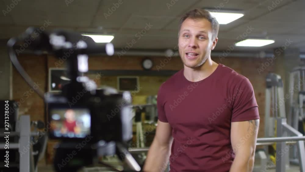 Cheerful gym coach waving, showing bicep and speaking on camera while recording video lesson for social media channel Cheerful gym coach waving, showing bicep and speaking on camera while recording video lesson for social media channel