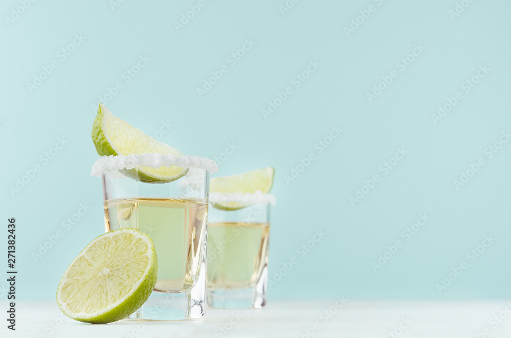 Obraz premium Gold tequila shots with salty rim and fresh lime slices on white table in modern elegant pastel blue interior, mexican cocktail with copy space for bar menu.