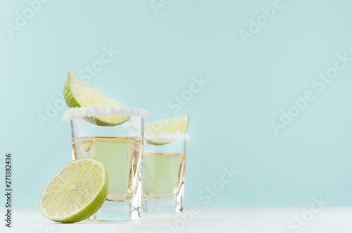 Cuadro en lienzo Gold tequila shots with salty rim and fresh lime slices on white table in modern elegant pastel blue interior, mexican cocktail with copy space for bar menu