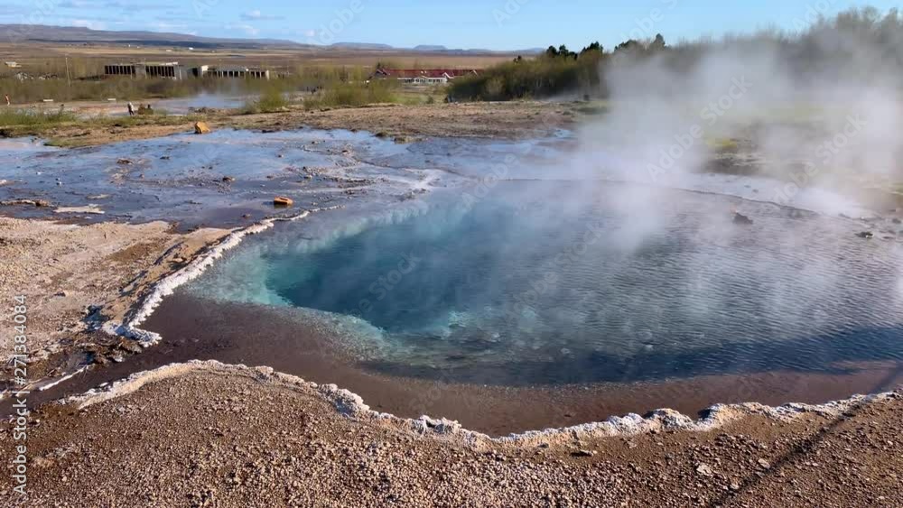 Steam coming from the beautiful turquoise water hot spring at the Strokkur geyser area in the southeast part of Iceland, surrounded by dry dirt landscape and mountains in the background Steam coming from the beautiful turquoise water hot spring at the Strokkur geyser area in the southeast part of Iceland, surrounded by dry dirt landscape and mountains in the background