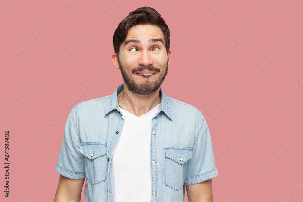 Portrait of crazy handsome bearded young man in blue casual style shirt standing with crossed eyes and looking with funny comedian face. indoor studio shot, isolated on pink background. Portrait of crazy handsome bearded young man in blue casual style shirt standing with crossed eyes and looking with funny comedian face. indoor studio shot, isolated on pink background.