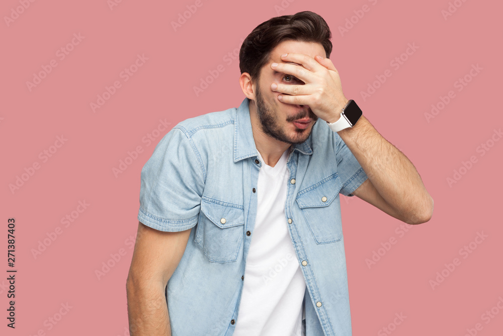 Portrait of shy, scared or peeking bearded young man in blue casual style shirt standing covering his eyes and looking at camera through his fingers. indoor studio shot, isolated on pink background. Portrait of shy, scared or peeking bearded young man in blue casual style shirt standing covering his eyes and looking at camera through his fingers. indoor studio shot, isolated on pink background.