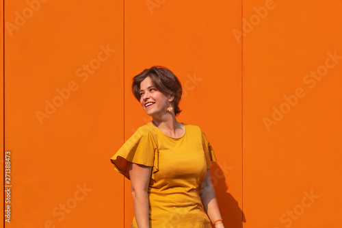 Attractive young smiling woman having fun in beautiful sunny day. Outdoor portrait of pretty brunette female model looking away of camera. Metal texture orange color wall on background. Head shot. 