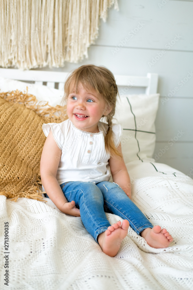 Fototapeta premium Beautiful little girl sitting on the bed in the bedroom. the baby has blonde hair and blue eyes. Jeans and white t-shirt