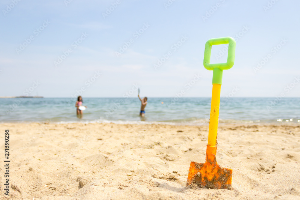 Yellow plastic toy shovel on sand beach at the seaside and kids playing in the sea in background. Yellow plastic toy shovel on sand beach at the seaside and kids playing in the sea in background.