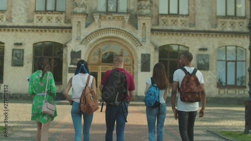 Back view of joyful multi ethnic friends walking towards college building in the morning for first lesson. Diverse multinational students going along campus alley to university together.