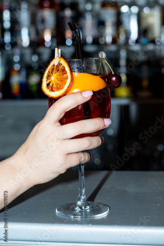 Alcoholic cocktail in a transparent glass on a thin leg. On the side of the chips are orange, two cherries on a branch, water drops on the glass. All on a blurred background. 