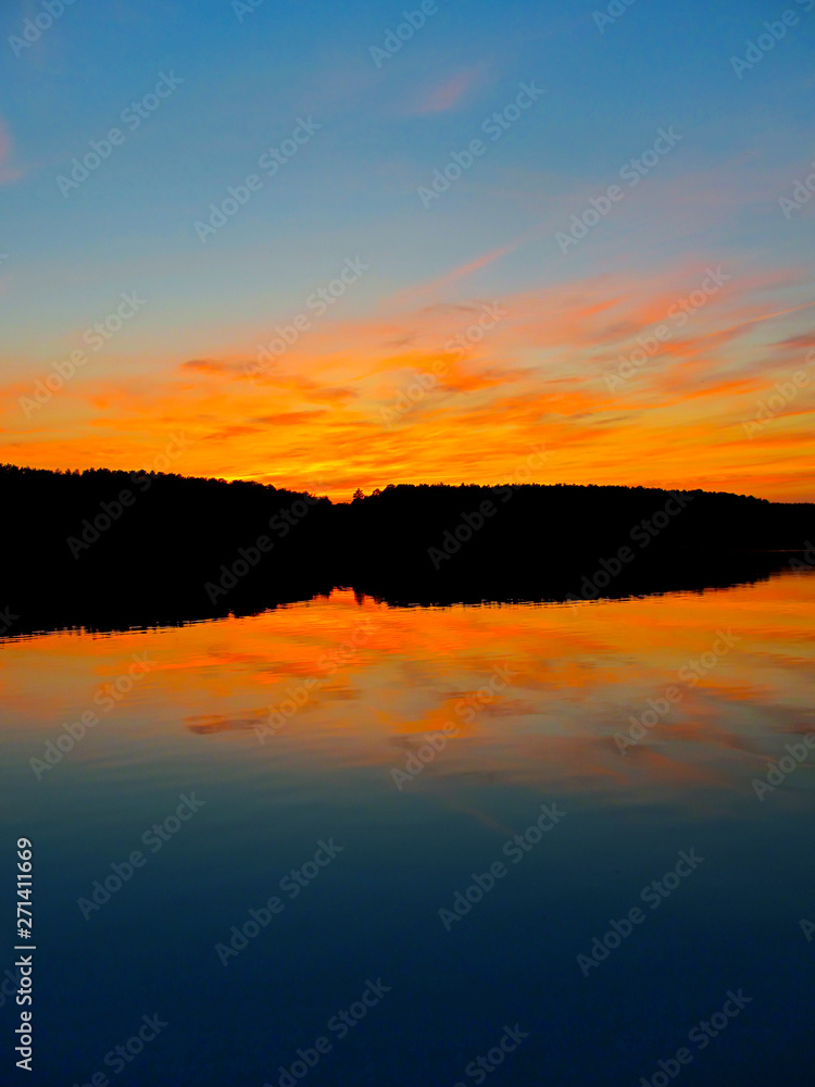 Bright colorful sunset over a lake. Beautiful reflection by a tranquil water of amazing sky and the lakeside at sundown in the summer evening. Lake Elovoe (Spruce Lake), South Ural, Russia.