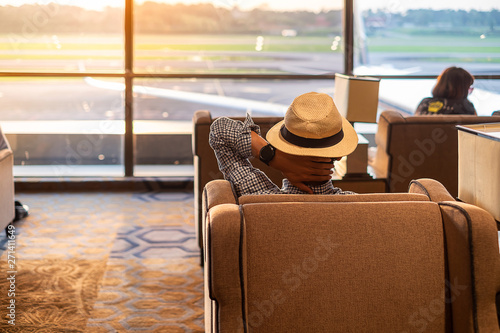 man traveler with hat looking to airplane in the morning sunrise, Asian passenger sitting and relax in modern lounge at international airport terminal. Travel concept