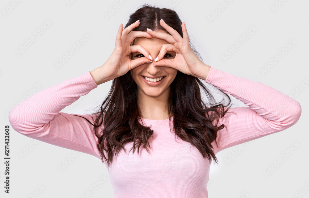Studio shot of beautiful young woman wearing pink casual outfit holding her hands at her eyes as if looking through binoculars or glasses. Happy female smailing against studio white wall background Studio shot of beautiful young woman wearing pink casual outfit holding her hands at her eyes as if looking through binoculars or glasses. Happy female smailing against studio white wall background