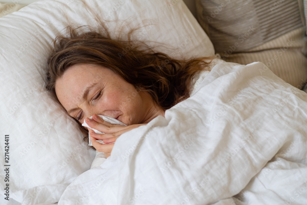 Sick young woman lying in bed covered with heavy blanket holding handkerchief Sick young woman lying in bed covered with heavy blanket holding handkerchief