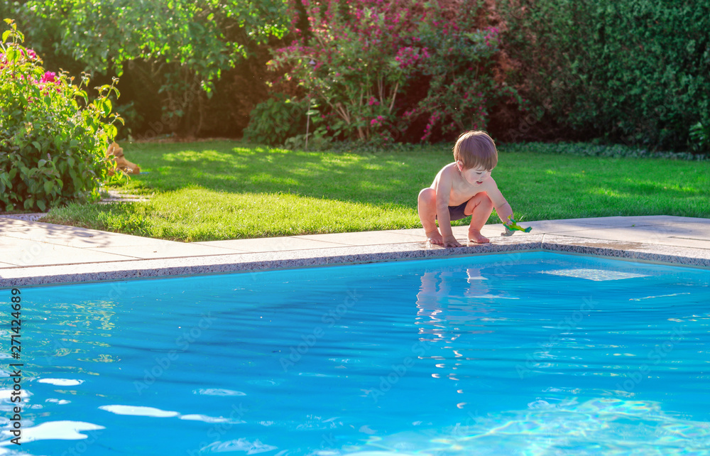 Little happy boy sitting on side of swimming pool in garden playing with his toy having fun. Summertime. Summer holidays.
