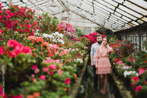 Young loving cheerful caucasian couple in glass greenhouse among colorful azalea flowers. Happy man and woman hugging and holding hands, spending time togehter.