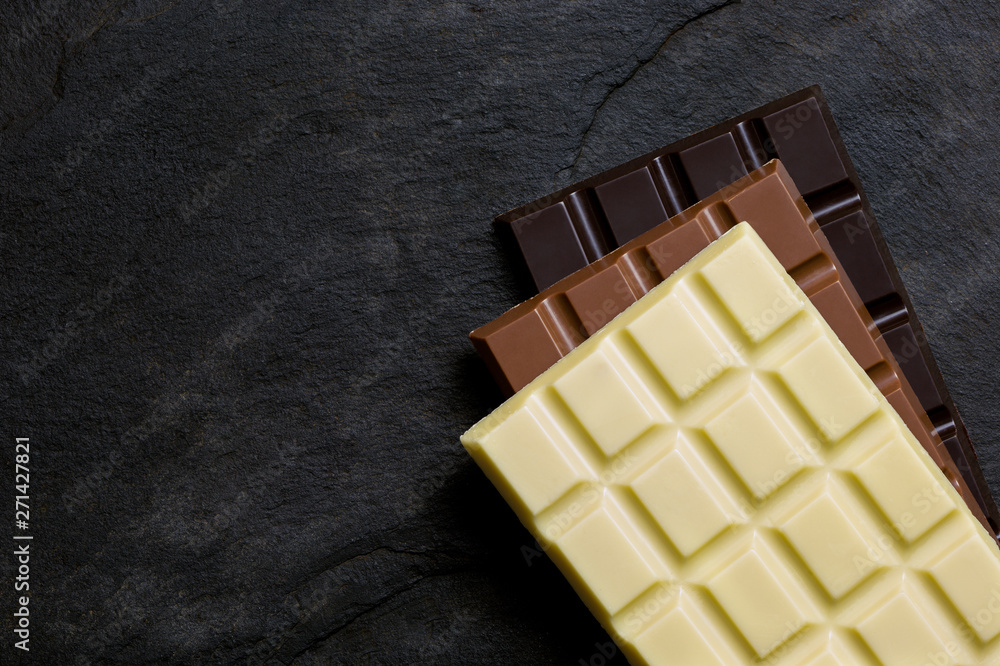 Slabs of white, milk and dark chocolate on top of each other on a black slate from above. Space for text. Slabs of white, milk and dark chocolate on top of each other on a black slate from above. Space for text.