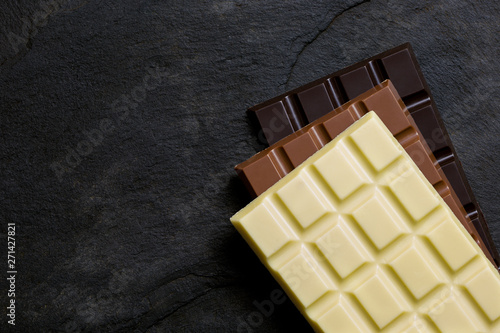 Obraz na plátně Slabs of white, milk and dark chocolate on top of each other on a black slate from above