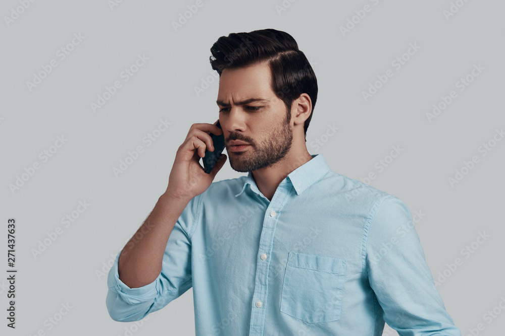 Bad news. Confused young man talking on the smart phone while standing against grey background Bad news. Confused young man talking on the smart phone while standing against grey background