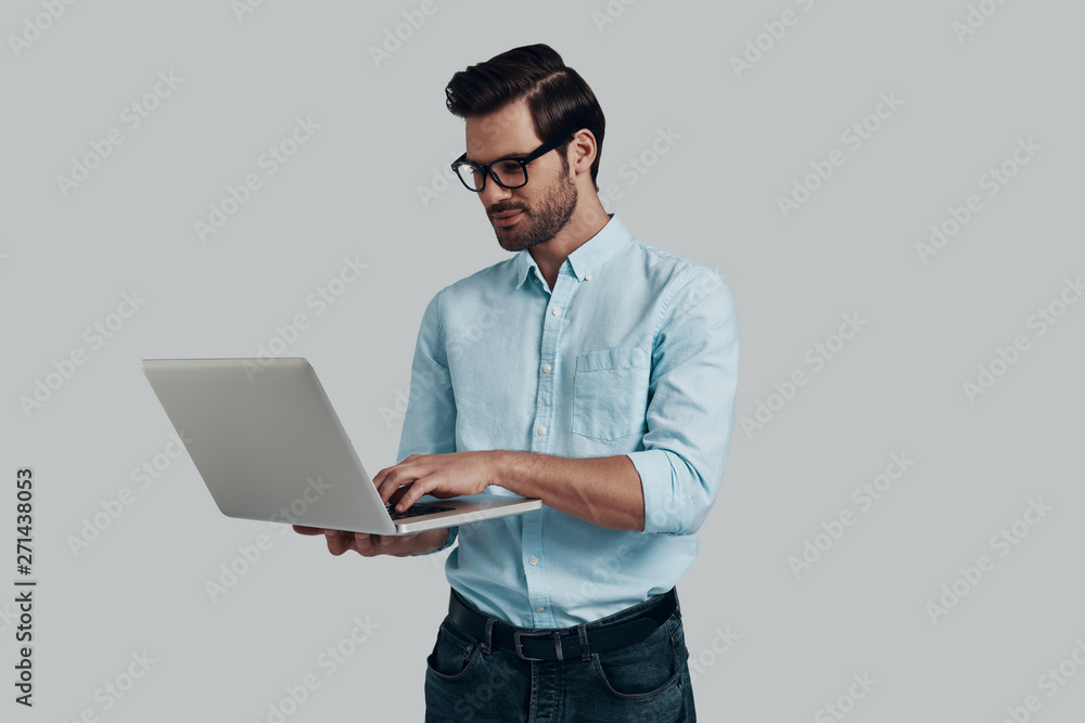 Paying attention to every detail. Young man using laptop while standing against grey background Paying attention to every detail. Young man using laptop while standing against grey background
