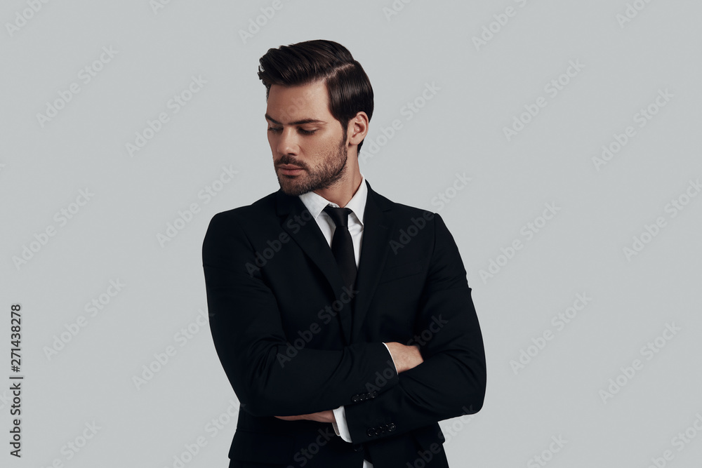 Used to look perfect. Handsome young man in full suit keeping arms crossed while standing against grey background Used to look perfect. Handsome young man in full suit keeping arms crossed while standing against grey background