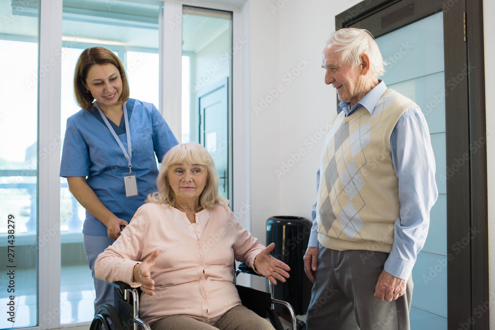Fototapeta premium senior woman in wheelchair talking with nurse in a hospital behind her standing senior husband.