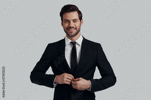 Always in style. Handsome young man in full suit adjusting jacket and looking at camera with smile while standing against grey background