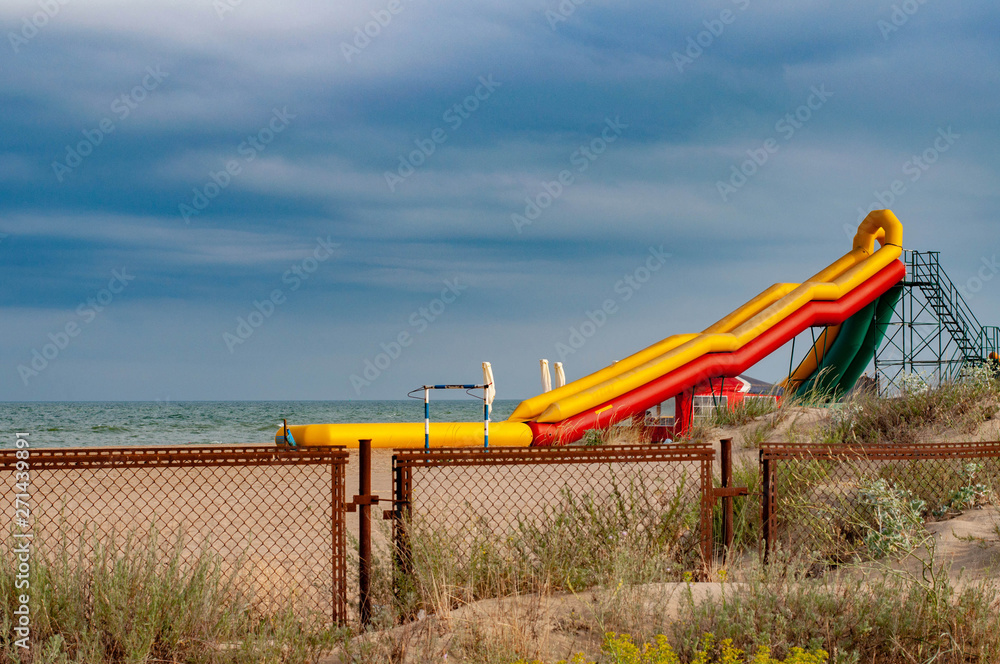 Colorful inflatable slide on beach sand of coastline. Seascape with rusty metal fence and aquapark equipment on blue sky background. Landscape with nobody and picturesque cloudscape. Summer vacation Colorful inflatable slide on beach sand of coastline. Seascape with rusty metal fence and aquapark equipment on blue sky background. Landscape with nobody and picturesque cloudscape. Summer vacation