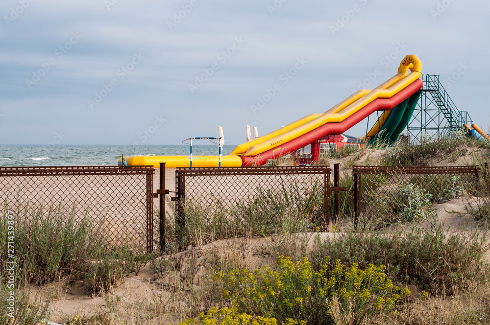 Zatoka, Odessa region / Ukraine - 06 27 2018: Yellow inflatable slide on beach sand. Seascape with rusty metal fence and aquapark equipment. Landscape with nobody in beginning of summer season Zatoka, Odessa region / Ukraine - 06 27 2018: Yellow inflatable slide on beach sand. Seascape with rusty metal fence and aquapark equipment. Landscape with nobody in beginning of summer season