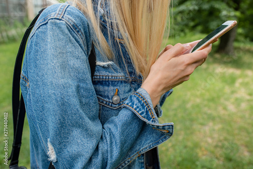 Young girl in a denim jacket and a smartphone in her hands. Life in social networks. Fashionable hipster. Trend concept. City women on the background of nature. Social modern problems. Lady blogger