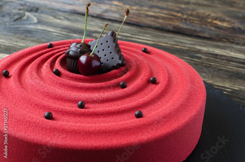 Contemporary cherry mousse cake covered with red velvet spray, decorated with chocolate elements and fresh cherries on black plate on wooden background