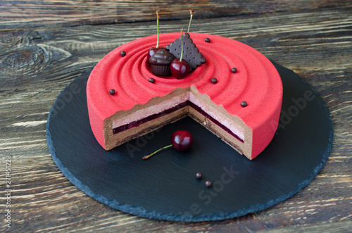 Part of contemporary cherry mousse cake covered with red velvet spray, decorated with chocolate elements and fresh cherries on black plate on wooden background