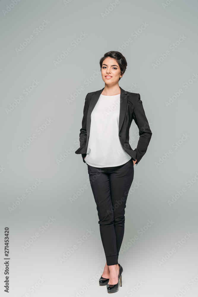 smiling mixed race businesswoman holding hands in pockets and looking at camera on grey background smiling mixed race businesswoman holding hands in pockets and looking at camera on grey background