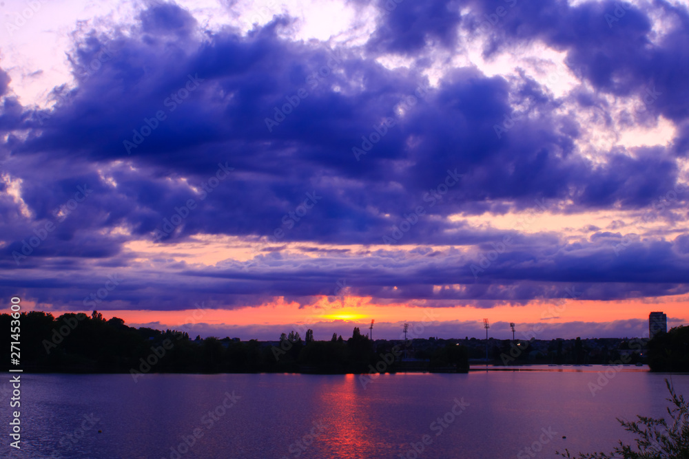 Awesome sunset over the city. Very colorful horizon with large cumulonimbus.  Dark sky with the rays of the sun piercing and reflected in the water of a lake