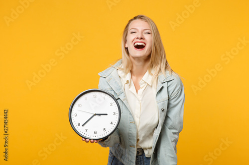 Portrait of laughing young woman in denim casual clothes looking camera, holding round clock isolated on bright yellow orange wall background in studio. People lifestyle concept. Mock up copy space.