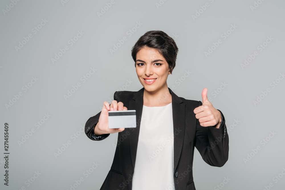 smiling mixed race businesswoman holding credit card and showing thumb up isolated on grey smiling mixed race businesswoman holding credit card and showing thumb up isolated on grey