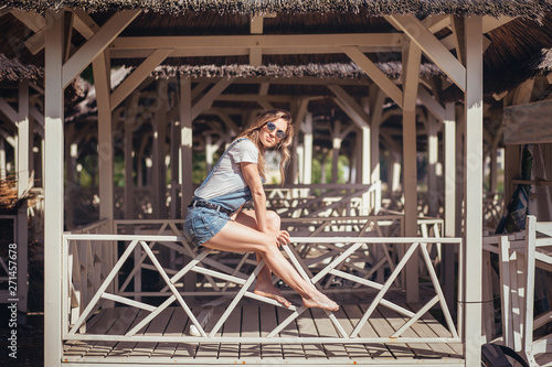 Positive caucasian happy young woman happily sunbathes in the sun and enjoys vacation sitting on a wooden thatched veranda by the sea on a sunny summer day during vacation