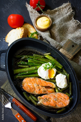 Salmon fish steak grilled with asparagus, poached egg in a frying pan on a rustic stone table. Healthy food.